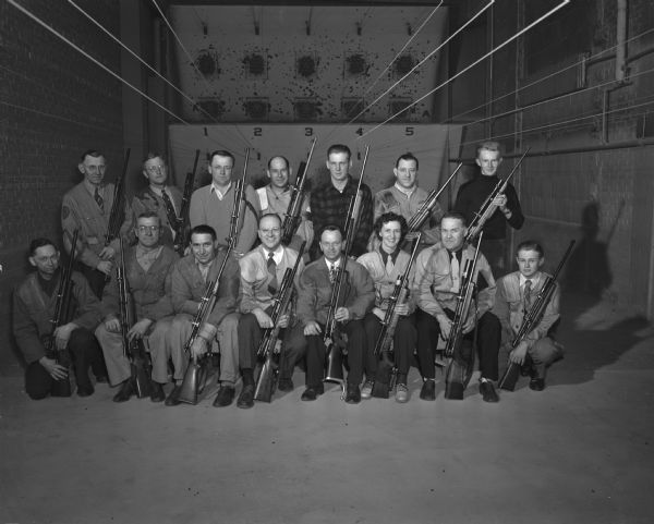 Fifteen men and women pose with their rifles in a indoor rifle shooting range.