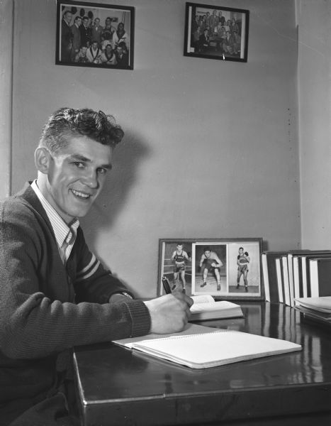 A man wearing a letter sweater sits at a desk.  A framed photograph of three boxers and several books stand on the desk and two framed photographs hang on the wall.