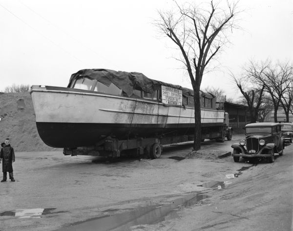 A large steel boat rests on a custom-built trailer pulled by a truck.  The boat is named "Skori" and is 18 tons in weight; 60 feet long; 12 feet wide and has seats for 90 people. The boat was built by the Cristy Corp., Sturgeon Bay and transported by Reynolds Transfer and Storage Co.  "The biggest vessel to be launched on the lake, the 'Skori' will be operated by Richard Butler, West high school teacher."