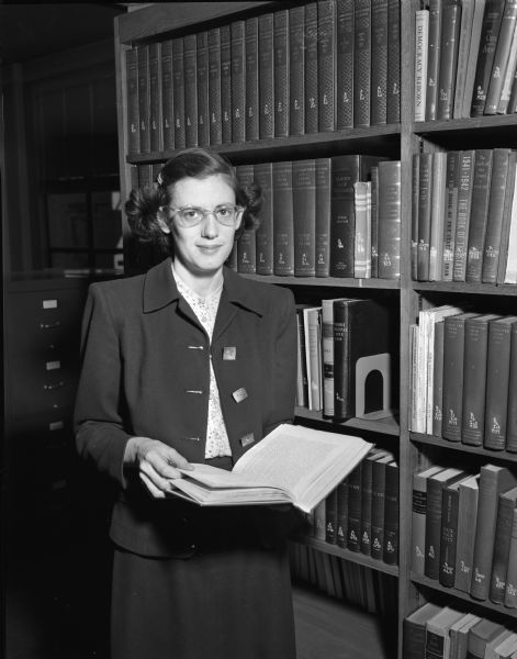 Portrait of Phoebe Hayes, librarian for the American Federation of State, County and Municipal Employees (AFSCME) and representative from the Madison Federal of Labor on the 1949 Madison Community Chest budget committee. She is holding a book and is standing next to a library shelf.