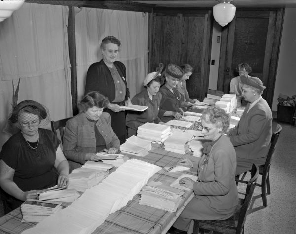 Madison volunteers stuffing envelopes for the financial drive of the American Cancer Society. Left to right: Mrs. Lewis W. (Leatha) Premo, cancer chairman of the Madison East Side Women's club; Mrs. Russell F. (Marjorie) Luxford, vice-commander of the Madison Cancer unit; Mrs. Charles E. (Alice) Hemingway, chairman of the Dane County unit, Wisconsin division of the society (standing); Mrs. Claude H. (Irene) Reading, Dane county radio chairman; Mrs. Sidney L. (Esther) Goldstine, chairman of exhibits of the Madison unit; Mrs. George Kahe, Dane county unit; Mrs. Ray (Adela) Baechler, Madison unit; Mrs. Joseph Berg, city unit, and Mrs. Joseph J.(Frances) Prokop, cancer chairman of the Catholic Women's club.