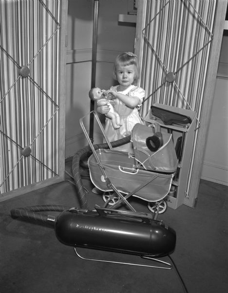 Barbara Jean Stoops, age 2, feeding her baby doll a bottle before trying out the new vacuum cleaner model at the Madison Home Appliance and Radio Show in the East High School gymnasium.