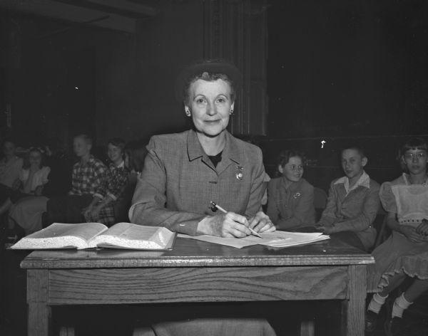 A woman, possibly a Badger Spelling Bee judge, sits at a desk with an open book, paper, and pencil.  A group of children sit behind her.