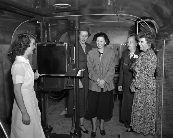 Miss Jeanne Gibbons (left), a technician with the Madison Board of Health, explaining the workings of the mobile X-ray unit to members of the Lakewood School District health committee where the unit is being used for a week. Maple Bluff Committee members, from left, are Mrs. Timothy Brown, Mrs. Robert O. McLean, Mrs. Russell A. Teckemeyer, and Mrs. Frederick L. Chapman.