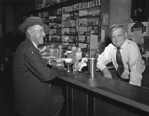 William Frazier, village president, sitting at a drug store counter visiting with druggist Burt Blank in the village of La Valle.
