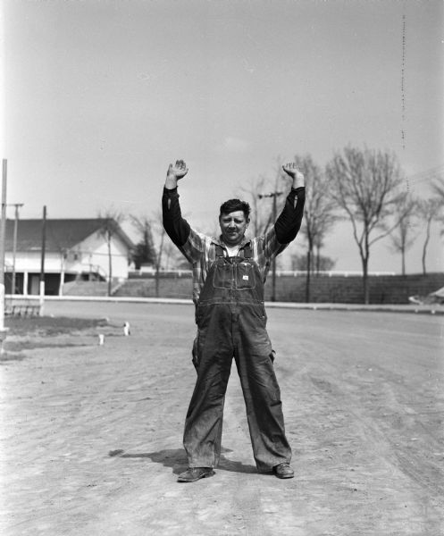 Clarence Zimbrich of Sun Prairie stands in the middle of a dirt road with his arms raised. He is a feed mill operator and vice-president of the Sun Prairie fire department, which sponsors the midget auto races held at Angell Park in Sun Prairie.