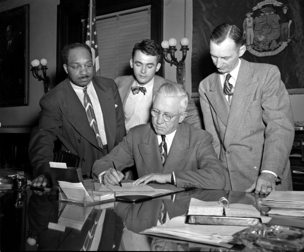 Governor Oscar Rennebohm, seated at his desk in the foreground, signs a bill into law ending segregation in the Wisconsin National Guard because of race, color, or creed. Standing behind the governor are three Democratic assemblymen, left to right: LeRoy J. Simmons and Charles J. Schhmidt, both from Milwaukee; and Thomas E. Taylor, Racine.