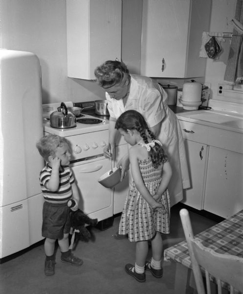 Mrs. Margaret Mentzner, a fulltime employee of the Family Service Agency, is shown caring and cooking for two small children whose mother is sick and absent.  The girl's hair is braided and tied with ribbon and the boy is holding a teddy bear.