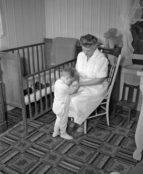 Mrs. Margaret Mentzner, a fulltime employee of the Family Service Agency, is shown hearing the bedtime prayers of a small boy whose mother is sick and absent. The boy is wearing pajamas and is standing next to his crib with his head nodded in prayer. His room has linoleum on the floor and is wallpapered.