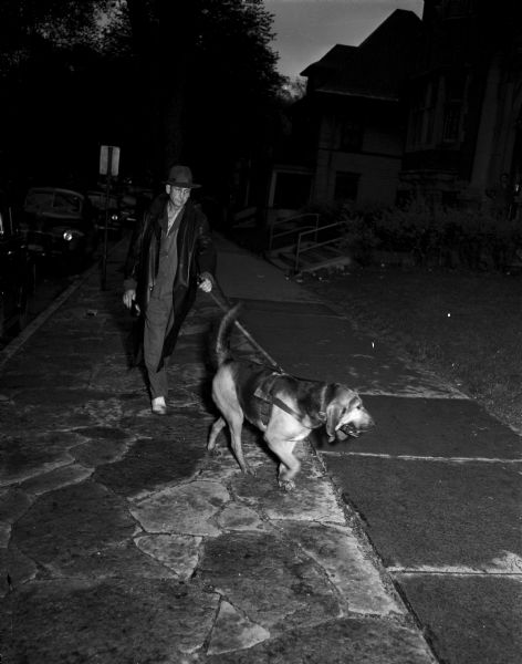 Ray Case, former County Traffic Officer, walks down a cobblestone walkway with his bloodhound, Bounding Bessie. The dog was used to trail an escaped jewel robber who took diamond rings from E.W. Parker, 9 West Main Street, on the Capitol Square.