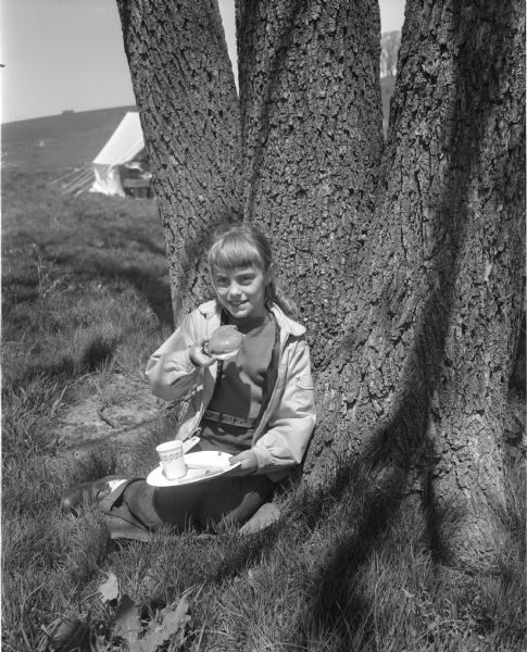 A girl sits on the grass next to some trees while eating a plate of food at a Girl Scout round-up.