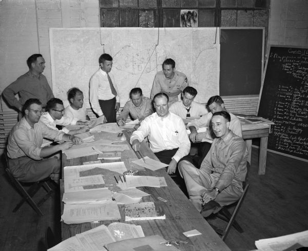 A group of eleven men gather around a table covered with newsprint notes.