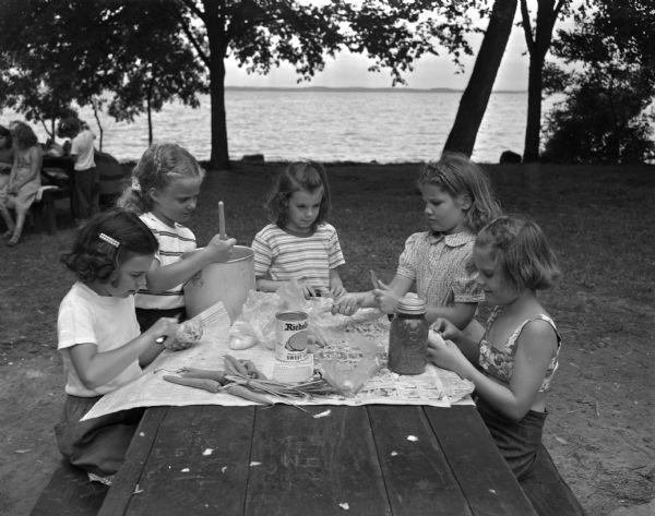 Four girls are gathered at a picnic table while making stew at Brownie Day Camp at Burrows Park. A lake is in the background. Left to right: Gloria Brandenstein, daughter of Mr. and Mrs. Leo Brandenstein, Sun Prairie; Sharon Householder, 2726 Myrtle Street; Irene Thompson, daughter of Mr. and Mrs. Lester Thomas, 1424 Jenifer Street; Jean Mickelson, daughter of Mr. and Mrs. Soren C. Mickelson, 2337 Hoard Street; and Donna Clements, daughter of Mr. and Mrs. Frank Clements, 2039 E. Johnson Street.