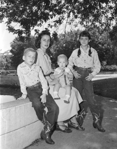 Mrs. Charles Beardsley of McAllen, Texas holds her son, Charles Jr., on her lap while sitting on a retaining wall at what appears to be a zoo or park.  Her other two children, King and Barbara, wear matching cowboy boots while standing on either side of her.  They are visiting Mr. & Mrs. A.G. Tappen (Mrs. Beardsley's parents) and Mr. & Mrs Charles Beardsley (Mr. Beardsley's parents).