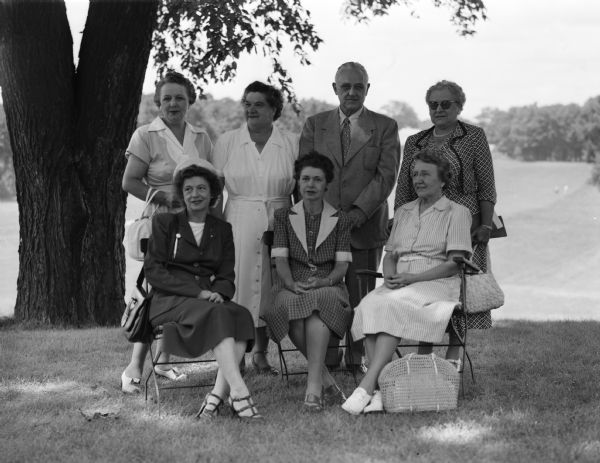 The tournament committee for the Wisconsin women's golf association tournament gathers beneath a tree for a group portrait. The tournament was to be held at the Maple Bluff Country Club. In the front row, left to right, are: Mrs. A.D. Kvool, Menomonee Falls North Hills Country Club, second vice-president of the WWGA and assistant tournament manager; Mrs. Walter P. Kimmel, Milwaukee Tripoli Golf Club, president of WWGA; and Mrs. E.J.B. Schubring, Madison Maple Bluff Country Club, a member of the rules committee. In the back row, left to right, are Mrs. Oscar T. Toebaas, Madison Maple Bluff Country Club, tournament chairman; Mrs. Ray H. Watson, Menomonee Falls North Hills Country Club, WWGA first vice-president and tournament manager; John St. John, president of the host club; and Mrs. C.J. Grootemaat, Milwaukee North Shore Country Club, WWGA treasurer and a member of the tournament committee.
