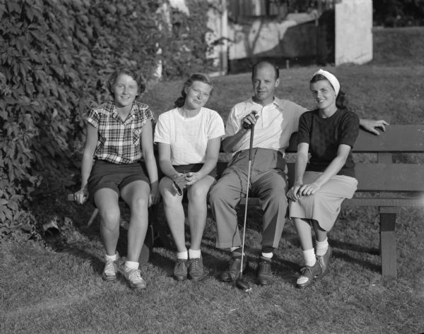 Three women and one man are sitting on a bench at Maple Bluff Country Club during the Wisconsin Women's Golf Association Tournament. Two of the women are Joan Coffeen of Green Bay, and Mary McMillin Jacobs, also of Green Bay.