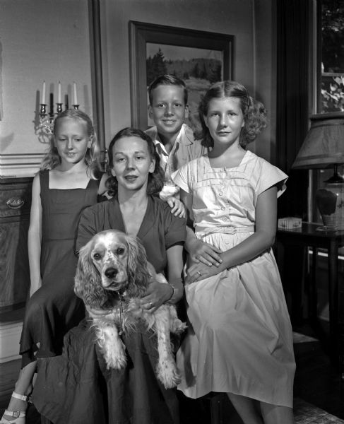 Group portrait of Mrs. Mark (Ruth) Schorer (center), her daughter, Suki, age 10 (at left), her son, Page, age 12 (standing behind his mother), and her niece, Nancy Gernon, age 11 (right), daughter of the Edward Taylor Gernons, 123 Kensington Drive, Maple Bluff. Mrs. Schorer is the daughter of Professor and Mrs. William H. Page, 515 North Carroll Street and is visiting from Berkeley, Caalifornia where her husband is a member of the faculty of the University of California. A dog, named Freckles, is being held by Mrs. Schorer.