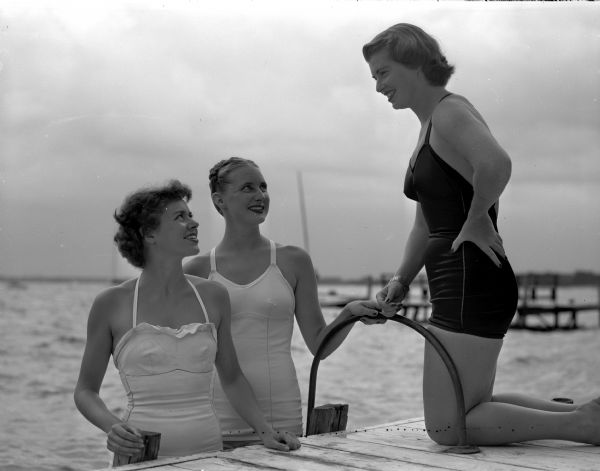 Three members of the University of Wisconsin water ballet team converse by a lakeside dock. Patricia Patterson, 260 Langdon Street, pictured at right, is the director of the ballet, and is shown discussing some of the water formations with Joan Donalds, St. Croix Falls, a sub-chairman for the event, at left; and Nancy Olmsted, Oshkosh, assistant director, at center. The event was held to promote the university's summer session prom.