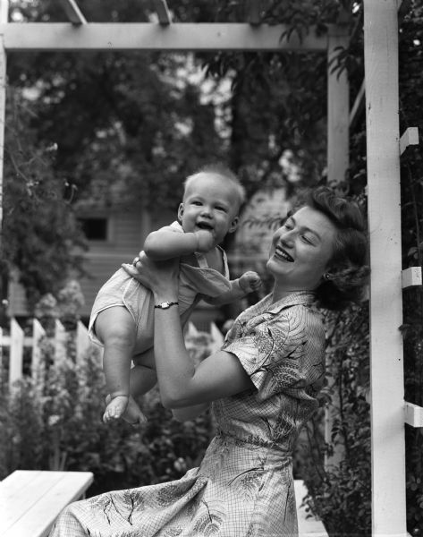 Mrs. Chester (Nancy) Rieck of Oak Park, Illinois holds her six-month-old son, Jeffrey Allan Rieck, while seated outdoors beneath a trellis. The Reicks are visiting Mrs. Reick's parents, Mr. and Mrs. Dwight (Lois) Fowler, 2211 Keyes Avenue.