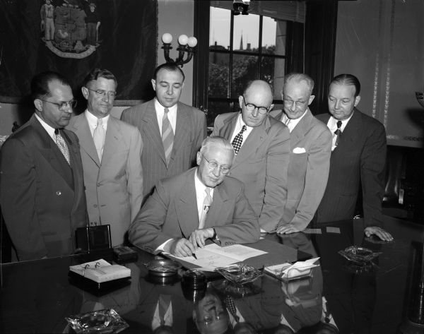 Governor Rennebohm signing an appropriation bill for $1,000,000 that will fund 94 added positions at the state's mental institutions. Watching him sign are, left to right: Harvey A. Stevens, Superintendent Southern Colony and Training School; A.J. Bayley, Director of State Dept. of Public Welfare; W.J. Urben, Superintendent Mendota State Hospital; B. Hughes, Superintendent of Winnebago State Hospital; Hobart R. Hunter, Superintendent Northern Colony and Training School; and J. Klepfer, Superintendent Central State Hospital.