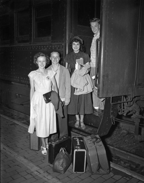 Four Madison high school youth are standing with their luggage near the entrance to a train car as they prepare to depart for the Red Cross training center at Camp Clear Lake near Battle Creek, Michigan. They are, left to right: Lois Brustman, Wisconsin High; Stefan Anderson, East High; Sue Lentz, West High; and Donald Noel, Central High. The training will be in the philosophy, purpose, program, and organization of the Junior Red Cross.