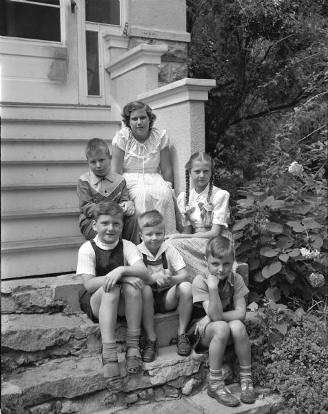 Four Latvian refugee children sitting  on the steps of their host family, the Allan Dicksons, 5435 Egan Court. In the front row, left to right, are Gundar and Vidvud Valdmanis and their cousin, Agnar. Behind them are John and Peggy Dickson and, on the right, Mara Valdmanis.