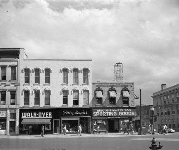 Refurbished store fronts along West Main Street at the corner of South Carroll Street. The building occupied by Walk-Over Shoe Store and Forbes-Meagher Music Company was lowered one story and given a shining new coat of cream-white paint. The Blied Building, housing Wisconsin-Felton Sporting Goods, was lowered two stories for safety purposes.