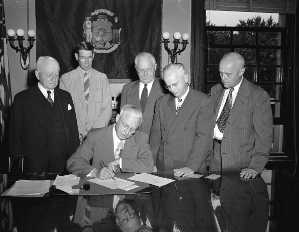 Governor Rennebohm signs a bill which will provide the University of Wisconsin with a new library. He is using a pen once used by President John Bascom. The pen and copy of the bill will be placed in a cornerstone of the new building. Looking on are Frank Sensenbrenner, Neenah, president of the University Board of Regents; University Librarian Gilbert Doane; Prof. Paul Knaplund; Ira H. Baldwin, University Vice-President, and Dean Mark Ingraham of the College of Letters and Science.