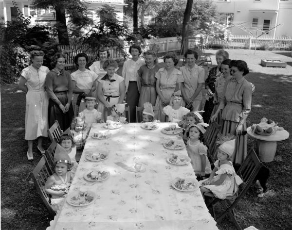 Ten children wearing birthday hats are seated around a  table.  Twelve women are standing in the background. Photo taken for Findorff.