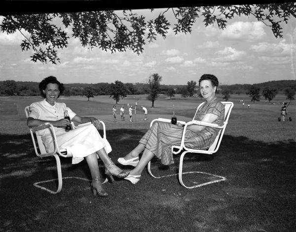 Mrs. Gilbert (Claudia) Stephan, 2205 West Lawn Avenue, member of the Nakoma Golf Club, and her guest for the club's annual Women's Guest Day, Mrs. John (Margaret) Stephan, 2264 Monroe Street, member of Maple Bluff Golf Club, are sitting beneath the shade of trees while having drinks. Mrs. John Stephan is Mrs. Gilbert Stephan's sister-in-law.