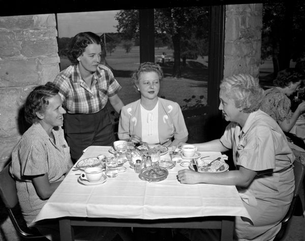 Standing and sitting around a table on the porch of the Nakoma Golf Club are club members (left to right): Mrs. Lester C. (Eleanor) Lee, 4229 Wanda Place, Mrs. Carl A. (Catherine) Felly, 814 Lewis Court, Mrs. William (Eunice) Schneller, 4226 Wanda Place, and Mrs. Frank (Melinda) Mazanet, 2533 Van Hise Avenue. The four women are attending the buffet luncheon that followed the club's Women's Organization Guest Day.