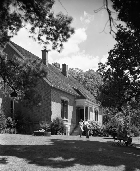 People are sitting on a bench and standing on the lawn surrounding the summer house on Governor Dewey's estate, called Stonefield.