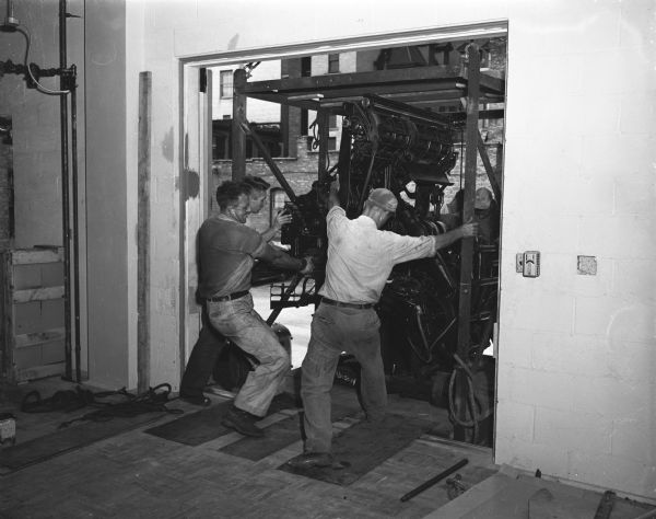 Three men move a Capital Times linotype machine into the new addition to the Wisconsin State Journal building, 115 South Carroll Street. The combined structures will become the plant of Madison Newspapers, Inc., and will house business and advertising offices, mechanical department and press-rooms, editorial offices and mailing room for two Madison newspapers - the Capital Times and the Wisconsin State Journal.