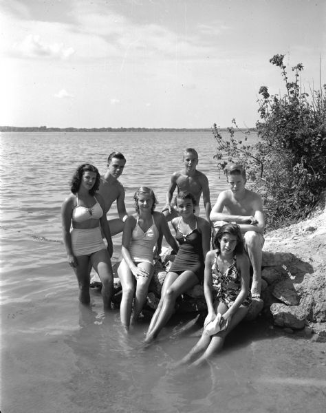 Group portrait of six members of the Shorewood Hills swim team, winners of the annual Madison city swimming meet. The girls, sitting on the rock, from left, are: Jane Gregg, Nancy Blume, and Judy Hicks. The boys, from left, are: Bruce Mohs, Jeff Blume, and Joe Irwin.  Also shown, to the extreme left, is Maple Bluff swimmer Barbara Ford who placed first in diving and third in free style.