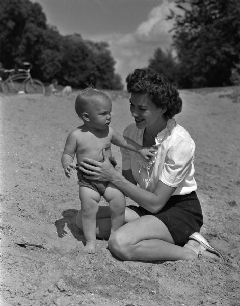 Mrs. Preston E. "Sandy" McNall Jr. holds her young son, Bruce, while on a beach. The McNalls are from West Lafayette, Indiana and are spending the summer at the home of Mr. McNall's parents, Professor and Mrs. P.E. (Eugenia) McNall, 926 Spaight Street. Mr. McNall Jr. is doing research at the hydraulics laboratory at the University of Wisconsin.
