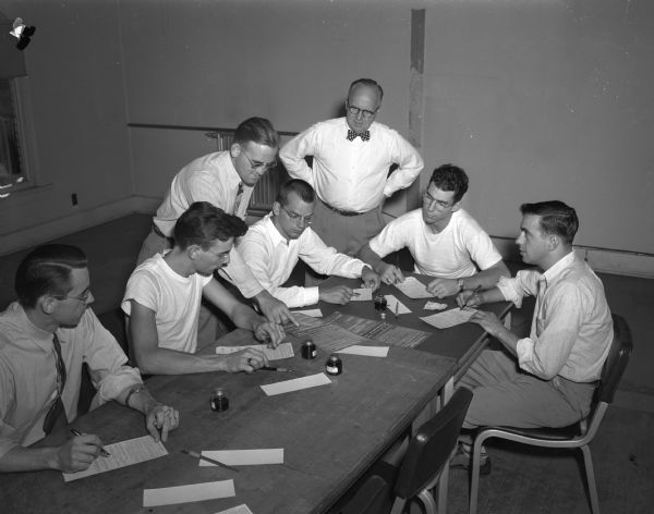 A group of veterans applies for insurance dividends at a table at the Veterans Administration office. Left to right are: Rolf N. Olsen, Madison; Ralph Cooper, West Allis; Wyndham Gary, Milwaukee; T.A. Cox, Texarkana, Texas; and Roger Murphy, Lancaster. Standing are John E. Tobin (left), senior contact representative for the Veterans Administration, and Eldon M. Stenjem, the office manager.