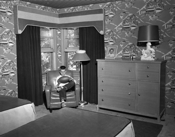 Bobby Fulton, 9 years old, son of Mr. and Mrs. Edward R. Fulton, 3425 Blackhawk Avenue, is shown sitting in his bedroom. Designed for rough wear, the room is furnished in leather and light woods, with accessories of sturdy corduroy; rubber tile covers the floor.