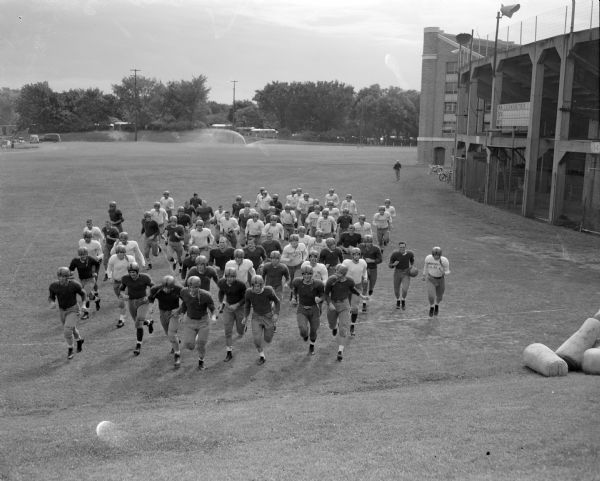 Elevated view of the 1949 University of Wisconsin football team, led by captain Robert "Red" Wilson, running onto the practice field outside Camp Randall Stadium for their first practice.