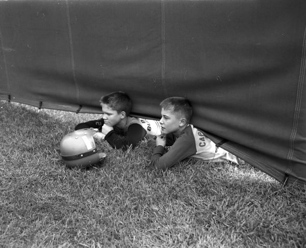 Two boys are lying on the ground to look under a canvas wall at the first practice of the University of Wisconsin 1949 football team outside Camp Randall Stadium. They are Pete Brandy (left) and Herman Herkert.