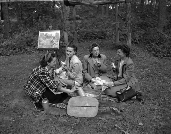 Madison Art Guild members sitting on a blanket for a picnic during an outdoor painting session. From left are Mrs. George W. Janet Washa, Mrs. Winfield (Ethel) Martin, Mrs. R.L. (Maude) Liebenberg, and Mrs. M.J. (Genevieve) Cass.