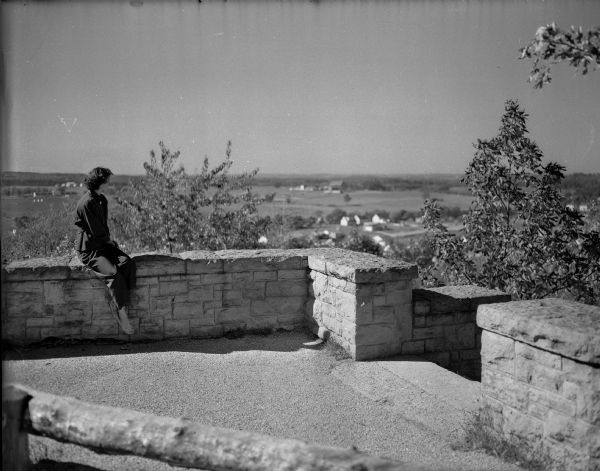 Mrs. Scott McGlasson, nee Shirley-Mae Riker, sitting on a stone wall at Sunset Point. She lives in Pomona, California, and is visiting her family in Madison.