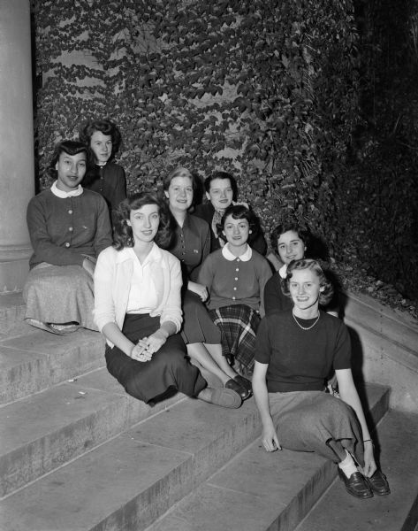 Seven Madison girls sitting on the steps to an ivy-colored building with the active president of the Omicron chapter of the Zeta Phi Eta national speech arts sorority. They are promoting the sorority's Founders' Day celebration to raise money for scholarships. From left are: Mary Majestic, Patricia Salisbury, Dolores Simms, chapter president Sandra Stekl, Thelma Rubin, Nadine Schuster, Mrs. John T. Reynolds, and Colette Slightam.