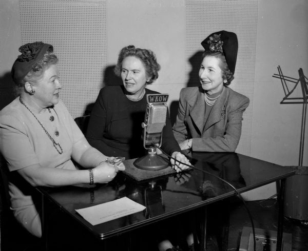 Three women discussing the aims and accomplishments of the Madison Business and Professional Women's Club being broadcast on the WKOW "Woman's World" broadcast program as part of an observance of National Business Women's Week. Left to right: Mrs. George R. Holhusen, co-chairman of the BPW radio and television committee; Mrs. W.E. (Luella) Mortenson, program director; and Mrs. Lois Allen Hart, radio and television committee chairman. The women are seated around a WKOW microphone on the table.