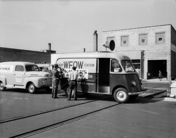Two men are installing a KAYSER'S banner on the side of the WFOW radio station truck for the Kayser safety parade on East Washington Avenue. The parade is part of a $100,000 Car Safety Contest, during which any automobile, regardless of make, is eligible for a free safety check at any Ford dealer.