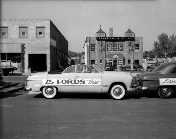 A person is sitting in the driver's seat of a Ford convertible in the foreground, with a banner that reads: "25 FORDS Free" on the side. In the background is the front of the Pennsylvania Oil Company (736 East Washington Avenue). The parade is part of a $100,000 Car Safety Contest, during which any car, regardless of make, is eligible for a free safety check at any Ford dealer.