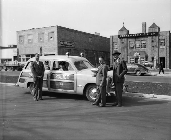 Three men are standing by a Dane County Police Ford station wagon during the Kayser Safety Parade in the 700 block of East Washington Avenue. Businesses in the background are Pennsylvania Oil Company and Hoffer Glass Company. The parade is part of a $100,000 Car Safety Contest, during which any car, regardless of make, is eligible for a free safety check at any Ford dealer.