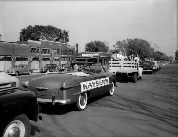 Line-up of Ford cars and trucks for the Kayser safety parade in the 700 block of East Washington Avenue. Musicians are in the back of a truck and commercial signs are in the background for Caterpillar, Pontiac Service and Chrysler. The parade is part of a $100,000 Car Safety Contest during which any car, regardless of make, is eligible for a free safety check at any Ford dealer.