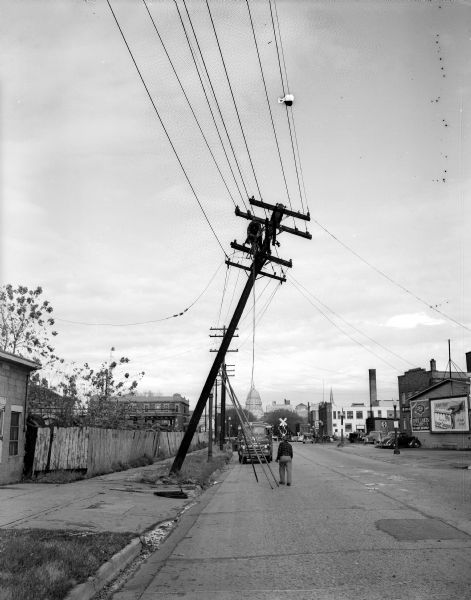 Workers repair an electrical pole that was partially blown over during a wind storm. The photograph was taken at the intersection of Regent Street and West Washington Avenue. Hotel Washington is visible left of center near the railroad crossing.