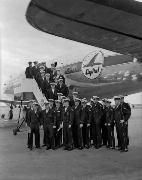 The Navy Midshipmen football team standing in front of and on the stairs to the airplane that brought them from Washington D. C. for the football game with the University of Wisconsin at Camp Randall.
