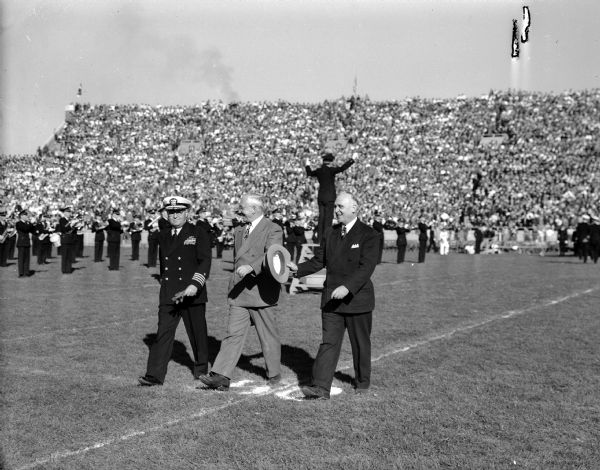 Governor Rennebohm between Captain Robert Pirie and University of Wisconsin President E.B. Fred on the field at Camp Randall at halftime of the Wisconsin - Navy football game. The Wisconsin marching band can be seen on the field, and fans are in the stands.